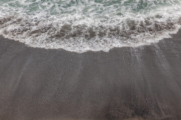 Waterscape background. Black sand beach with white milky foam waves. Nature and environment concept. Black and white contrast. Daylight. Copy space. Volcanic beach in Bali