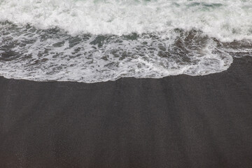 Waterscape background. Black sand beach with white milky foam waves. Nature and environment concept. Black and white contrast. Daylight. Copy space. Volcanic beach in Bali