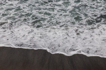 Waterscape background. Black sand beach with white milky foam waves. Nature and environment concept. Black and white contrast. Daylight. Copy space. Volcanic beach in Bali