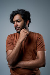 Indian man with beard in orange shirt poses in gray background looking away with hand under his chin.