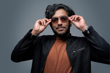 Obraz premium Stylish hindu hipster wearing fashionable clothing poses in gray background taking off his sunglasses.