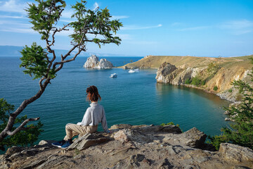 Fototapeta premium Girl with dreadlocks sits on rock under pine tree and looks at bay. Summer European view. Travel concept
