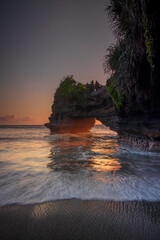 Natural arch. Batu Bolong temple on the rock during sunset. Seascape background. Motion milky waves on black sand beach. Copy space. Vertical layout. Tanah Lot, Bali