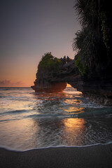 Fototapeta na wymiar Natural arch. Batu Bolong temple on the rock during sunset. Seascape background. Motion milky waves on black sand beach. Copy space. Vertical layout. Tanah Lot, Bali