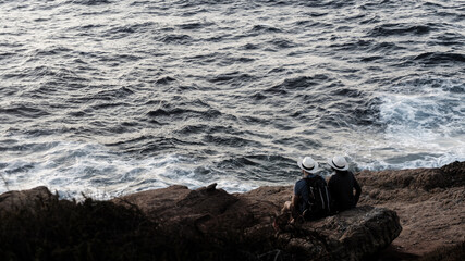 Young romantic couple in white identical hats with a backpack are watching the sunset while sitting on the rocky seashore.