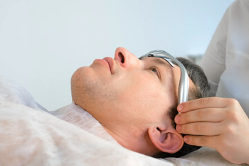 Chiropractor doctor is putting on electronic headband from migraine on man's forehead. Treatment of headaches with electric current in clinic on procedure with manual physiotherapist.