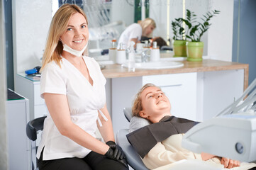 Cheerful female stomatologist smiling while young woman lying in dental chair. Dentist in sterile gloves sitting beside patient in dental office. Concept of dentistry and dental care.