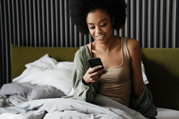 Happy african american girl using mobile phone while sitting in bed