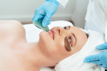 Beautiful woman having guasha scraper face massage done by cosmetologist