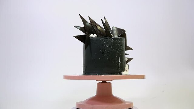 Black cake decorated with chocolate and marshmello slices rotates in a circle on a pink stand. Unusual stylish dessert on a white background. Space style cake.