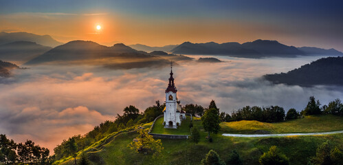Fototapeta premium Sebrelje, Slovenia - Aerial panoramic drone view of the beautiful hilltop church of St.Ivan (Sv. Ivan Cerkev) at sunrise with huge morning fog bellow the valley and Julian Alps at background