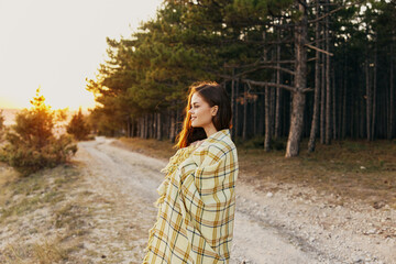 Happy woman tourist with a plaid on her shoulders stands on the road near high fir trees