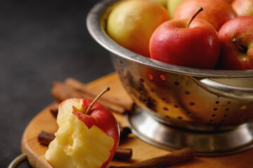 Bitten apple on cutting board with scattered cinnamon sticks on metallic bowl full of red ripe...