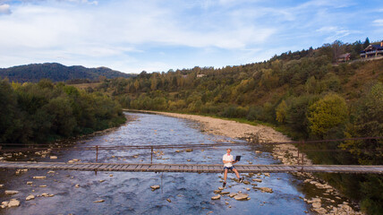 Aerial drone view of young man in white shirt sitting on wooden bridge over mountains river and using laptop. Success freelancer 
