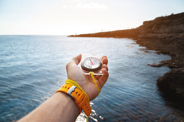 A man's hand with a wristwatch bracelet holds a magnetic compass against the background of the sea surface and the coast © yanik88