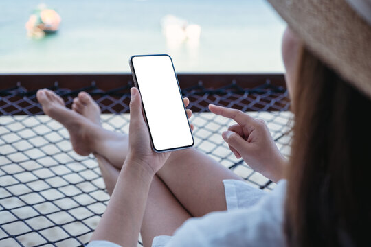 Mockup image of a woman holding mobile phone with blank desktop screen while lying down on hammock on the beach