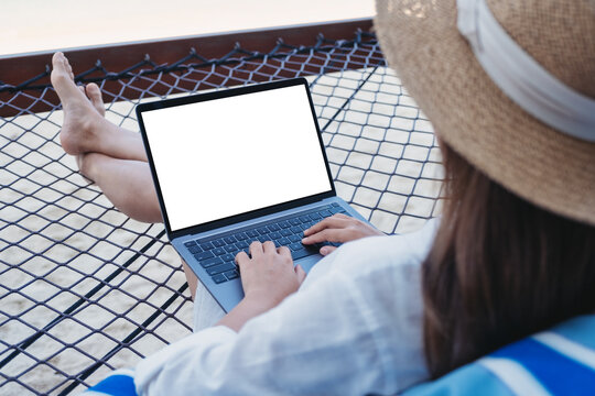 Mockup image of a woman using and typing on laptop computer with blank desktop screen while lying down on hammock on the beach