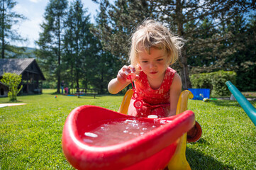 Cute blonde girl playing outdoor with water in plastic wheelbarrow.