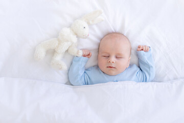 baby boy sleeping on the bed lying on his back with a stuffed toy hares in blue pajamas hands up, healthy newborn sleep