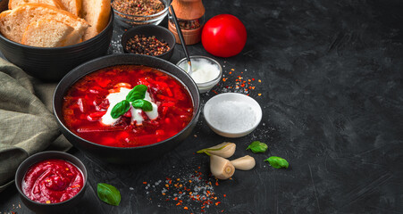 Fresh, delicious borscht in a deep black plate, on a background of bread, spices, garlic and sour cream on a black background.