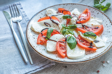 Caprese salad with sliced fresh tomatoes, mozzarella cheese and basil served on a white plate on light gray table surface.