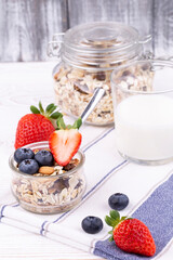 Healthy breakfast with granola and milk, fresh strawberries and blueberries on a white wooden table in rustic style, close up, vertical.