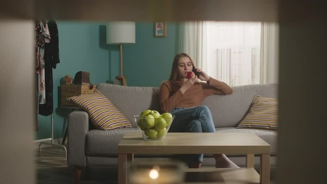 Young woman in blue jeans and brown sweater sits on beige sofa on window background, bites red apple and talks on phone. On coffee table is glass jar with green apples. Dolly in.