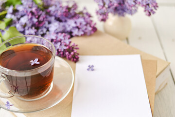 Romantic background with a cup of tea, lilac flowers and a book over a white wooden table. Leisure concept, spring breakfast. Mockup.