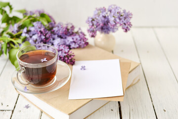Romantic background with a cup of tea, lilac flowers and a book over a white wooden table. Leisure concept, spring breakfast. Mockup.