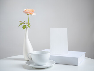 Mock up white  invitation card ,coffee cup and beautiful orange rose flowers in modern vase set on white wood table ,greeting card in soft white tone still life