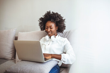 Happy young woman in Bluetooth headphones sit on couch study on laptop listening to music, smiling positive girl on sofa wear wireless earphones have skype or web conversation on computer