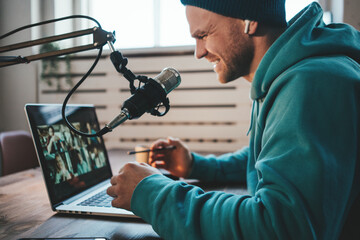 Handsome smiling host streaming his live podcast using microphone and laptop at his homemade...