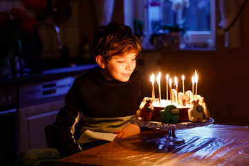 Adorable happy blond little kid boy celebrating his birthday. Child blowing candles on homemade baked cake, indoor. Birthday party for school children, family celebration