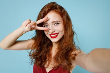 Close up young smiling readhead curly woman in red party evening dress gown doing selfie shot on mobile phone cover eye with victory v-sign gesture isolated on pastel blue background studio portrait.