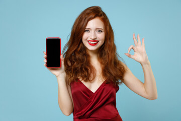 Young caucasian happy readhead curly woman 20s in red party dress gown hold mobile cell phone with blank screen workspace area show ok okay gesture isolated on pastel blue background studio portrait.