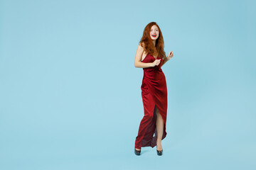 Full length of young caucasian smiling excited happy readhead curly attractive woman 20s wearing red party evening dress gown walking going isolated on pastel blue color background studio portrait