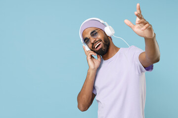 Young cool unshaven black african man 20s wear violet t-shirt hat glasses white headphones listen...