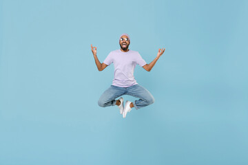 Fototapeta na wymiar Full length young unshaven black african man in violet t-shirt hat glasses hold spread hands in yoga om gesture relax meditate jump high levitate isolated on pastel blue background studio portrait.