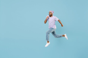 Fototapeta na wymiar Full length of young confident excited smiling happy unshaven black african man 20s in violet t-shirt hat glasses jump high run fast hurrying isolated on pastel blue color background studio portrait.