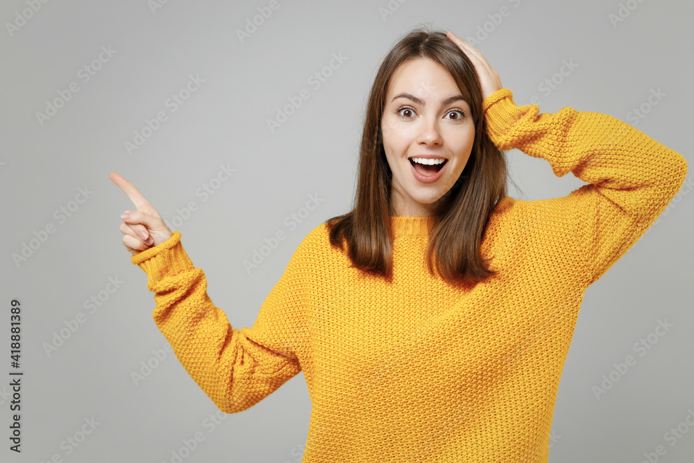 Wall mural Young excited happy surprised shocked caucasian woman in casual knitted yellow sweater point index finger aside on copy space area mock up holding head isolated on grey background studio portrait