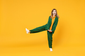 Full length little blonde kid happy girl 12-13 year old in casual clothes raised up leg look camera with intertwined fingers isolated on yellow orange background children Childhood lifestyle concept