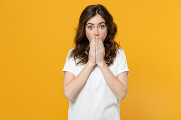 Young shocked impressed surprised scared eueropean caucasian student woman 20s wear white basic casual t-shirt cover mouth with hand look camera isolated on yellow orange background studio portrait