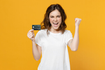 Young smiling happy rich caucasian student woman 20s in blank print design white basic t-shirt hold credit bank card do winner gesture clench fist isolated on yellow orange background studio portrait.