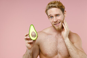 Smiling bearded naked young man 20s perfect skin hold half of fresh avocado fruit put hand on cheek...