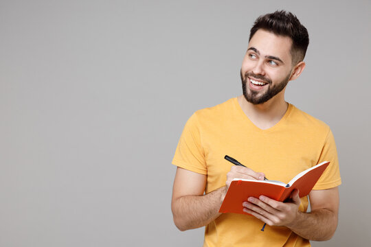 Young caucasian dreamful bearded attractive active happy student smart man 20s wear casual yellow basic t-shirt writing down in exercise book diary isolated on grey color background studio portrait
