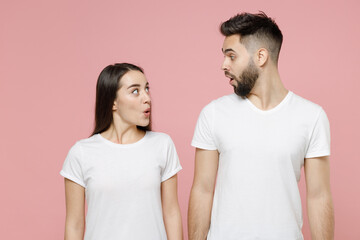 Young shocked fun couple two friends bearded man brunette woman in white basic blank print design t-shirts posing looks to each other say wow isolated on pastel pink color background studio portrait