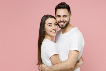 Young happy couple in love two friends bearded man brunette woman 20s in white basic t-shirts looking camera standing smiling hugging embrace isolated on pastel pink color background studio portrait.