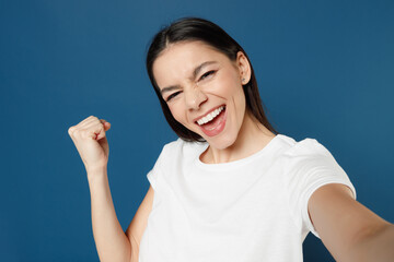 Fototapeta premium Young smiling overjoyed excited happy latin woman 20s in white casual basic t-shirt doing selfie shot on mobile phone do winner gesture clench fist isolated on dark blue background studio portrait.