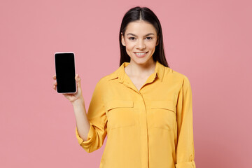 Young latin smiling positive happy cute attractive woman 20s in yellow shirt hold mobile cell phone blank screen workspace area looking camera isolated on pastel pink color background studio portrait