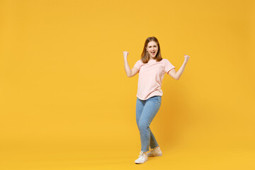 Fototapeta na wymiar Full length of young caucasian overjoyed excited woman 20s in basic pastel pink t-shirt, jeans do winner gesture clench fist celebrating say yes isolated on yellow color background studio portrait.